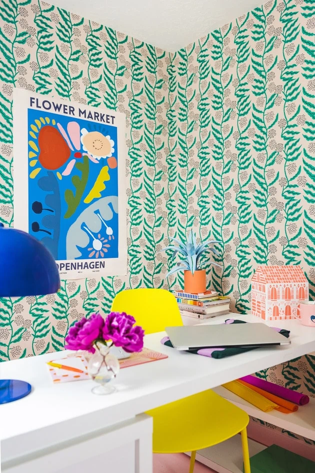 A brightly colored office. There's green floral wallpaper, a pink floor, a yellow chair, a white table, an orange paper house, a blue poster with colorful abstract flowers, and office supplies.
