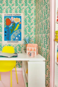 A brightly colored office. There's green floral wallpaper, a pink floor, a yellow chair, a white table, pink-painted doorframes, an orange paper house, a blue poster with colorful abstract flowers, and office supplies.