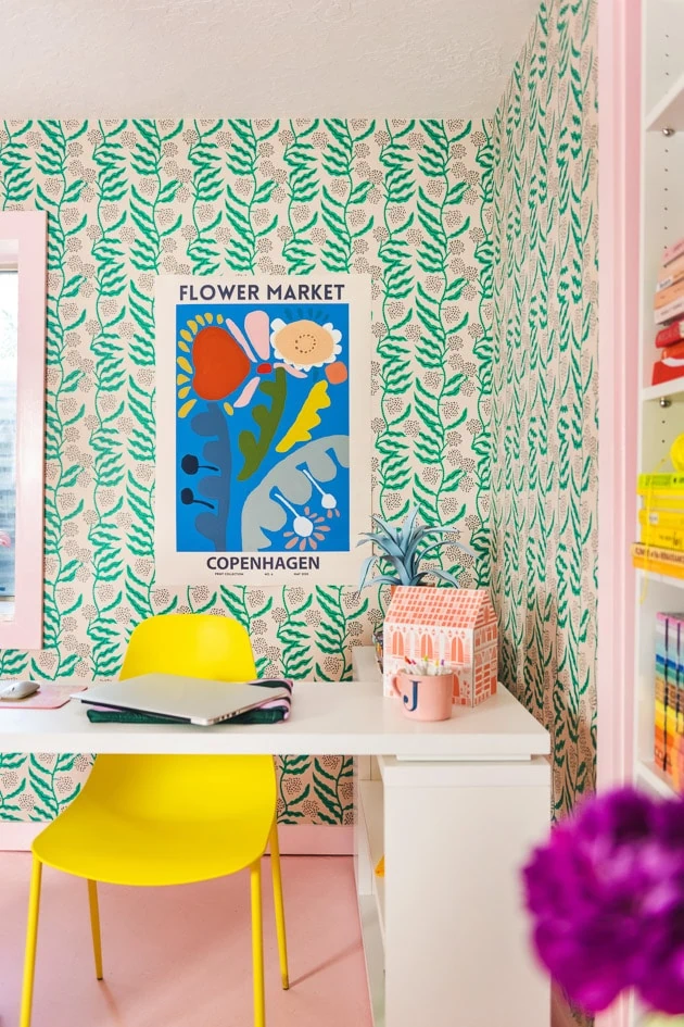 a blue poster with flowers drawn on it hangs on a green botanical wallpapered wall. There's a bright yellow chair, white office furniture, and a pink room in the space.