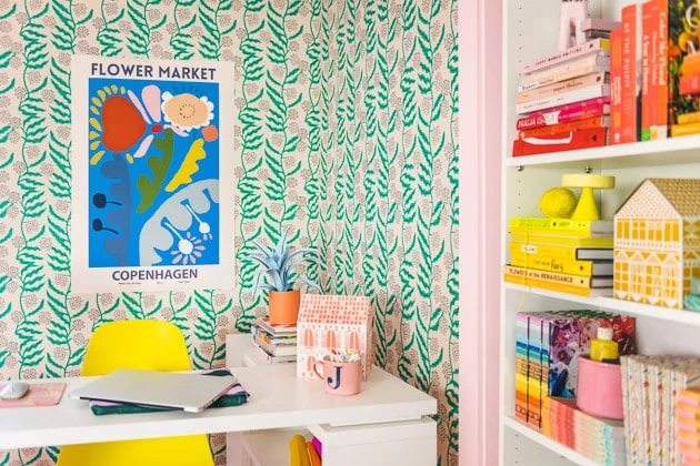Interior shot of the finished office. You can see the wallpaper, the blue art print with colorful flowers, and the desk with an orange paper house and yellow chair, and you can also see a shelf installed in the closet that's filled with a rainbow of books. 