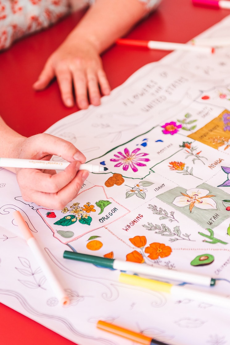 The coloring map is on a red table. Colorful markers are scattered around and some hands reach in to color it.