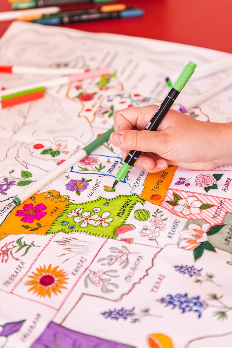 The coloring map is on a red table. Colorful markers are scattered around and some hands reach in to color it.