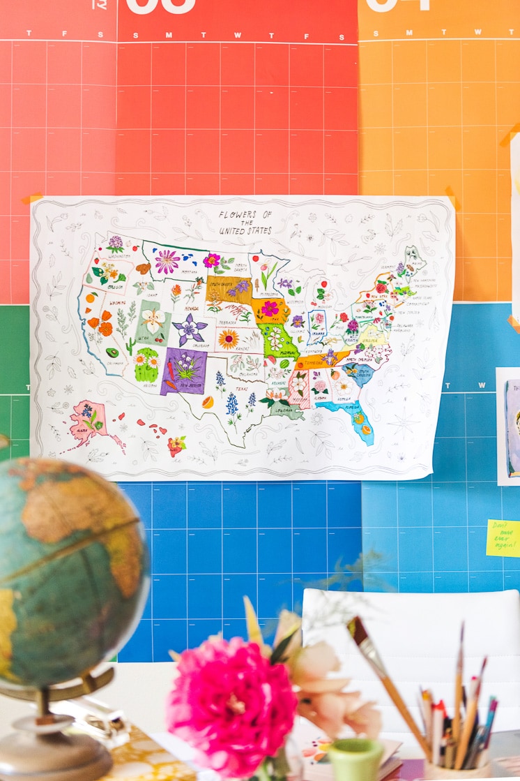 The State Flower Map Coloring Page hangs on Brittany's rainbow calendar with some art prints, a painting by Jasper, and a cutout bird. A colorful desk with flowers, a laptop, books, and a globe is in front of the calendar.