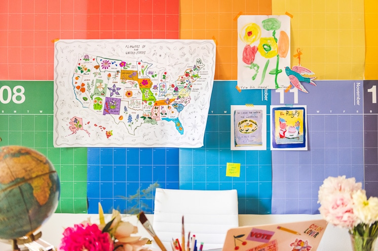 The State Flower Map Coloring Page hangs on Brittany's rainbow calendar with some art prints, a painting by Jasper, and a cutout bird. A colorful desk with flowers, a laptop, books, and a globe is in front of the calendar.