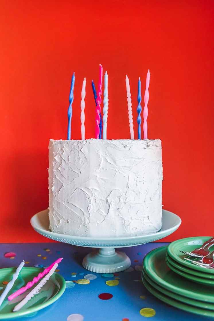 A birthday cake with twisted birthday candles on it. The background is red and the table is blue. There are candles, green plates, and some confetti around.
