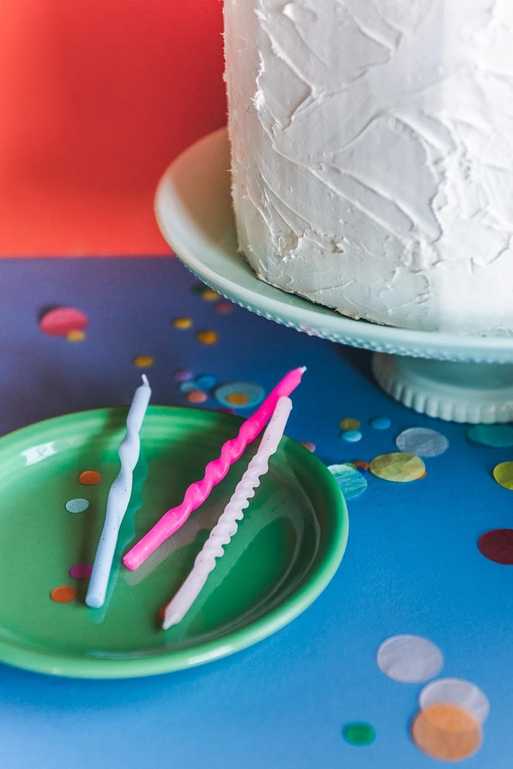 three twisted candles on a green ceramic plate next to a birthday cake. There's confetti around and the background is red and blue.