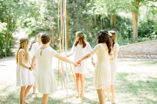 women dressed in white dance around a DIY maypole in a green park with dappled light. 