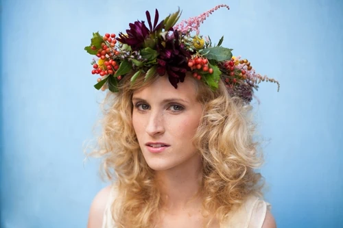 A woman wearing a lavish flower crown of dahlias and berries and greenery looks at the camera. The background is sky blue.