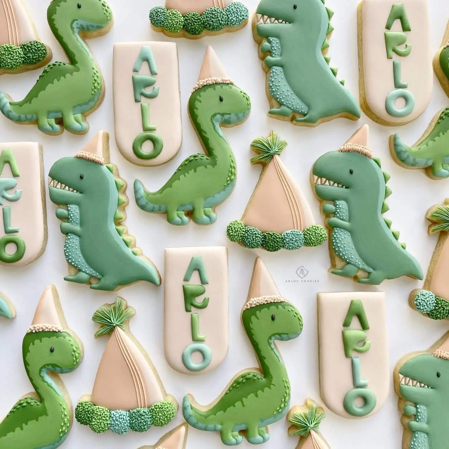 Decorated cookies in the shapes of dinosaurs, party hats, and pendants with the name "Arlo" arranged in a flat lay.
