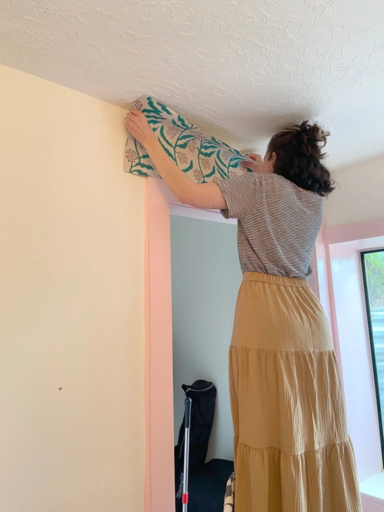 Hailey stands on a stepstool and installs the green vined wallpaper. She's wearing a striped t shirt and a tan skirt.