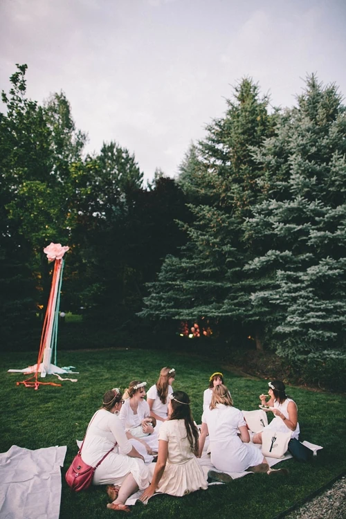 women dressed in white eat on a picnic blanket. In the background, a maypole stands in front of some pine trees. It's dusk.