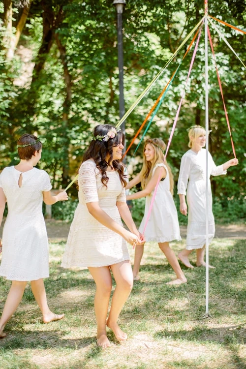 women dressed in white dance around a DIY maypole in a green park with dappled light.