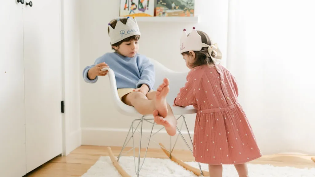 Two kids wearing linen birthday crowns and playing. A girl is wearing a pink dress and white crown, and a boy is wearing a blue sweater, yellow shorts, and a grey crown. They're in a light-filled room.