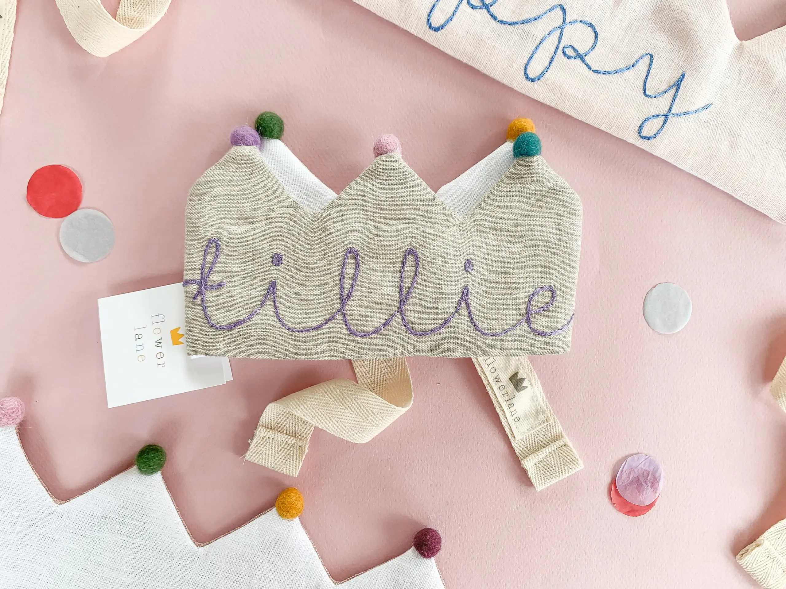 A grey linen kids birthday crown with rainbow pom poms and the name "tillie" embroidered on it. It's on a pink background. 