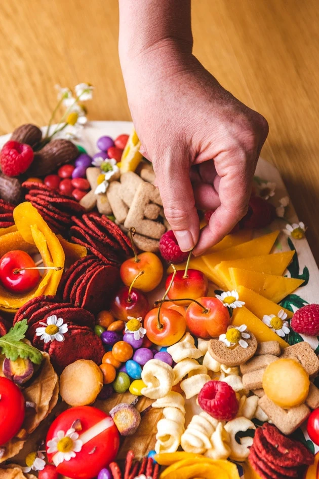 A hand reaches down to a charcuterie board laden with fruits, candies, snacks, veggies, cheese, and crackers. 