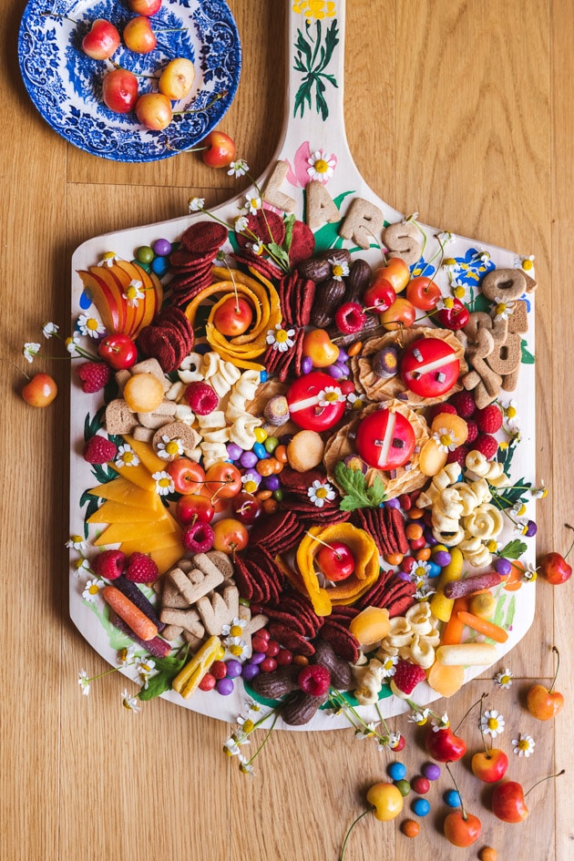 Painted charcuterie board laden with snacks and cookies and fruits and cheeses and treats. In the top left corner a bowl full of cherries spills over and in the bottom right corner some candies, cherries, and flowers are arranged. 