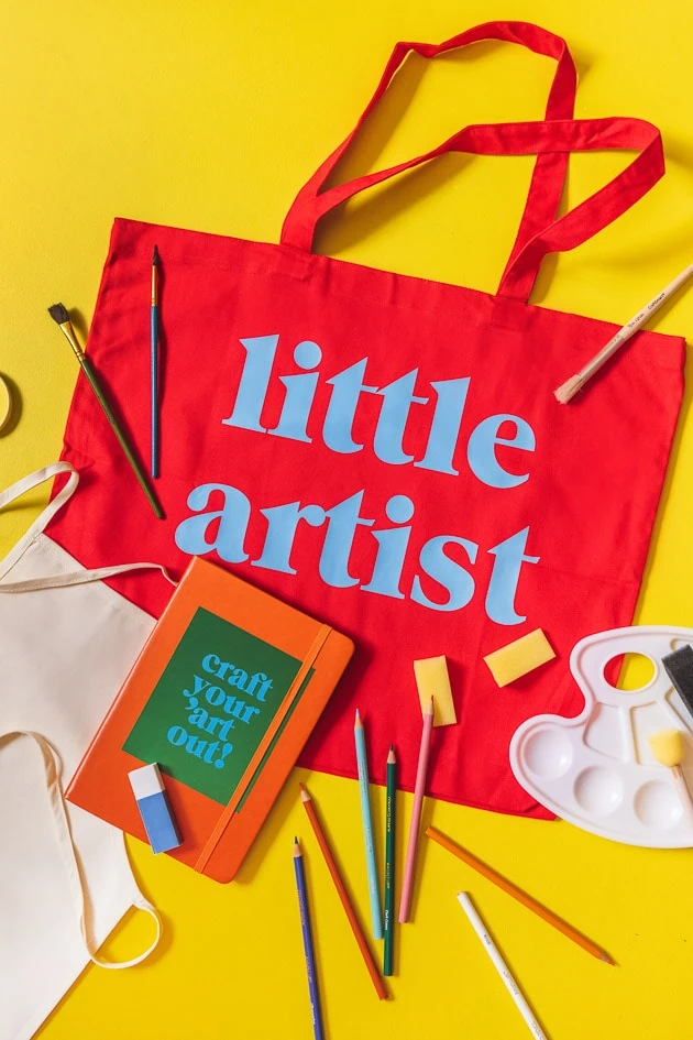Little Lars art kit, including a red tote, a paint palette, paintbrushes, colored pencils, a sketchbook with a sticker, an eraser, and an apron on a yellow background