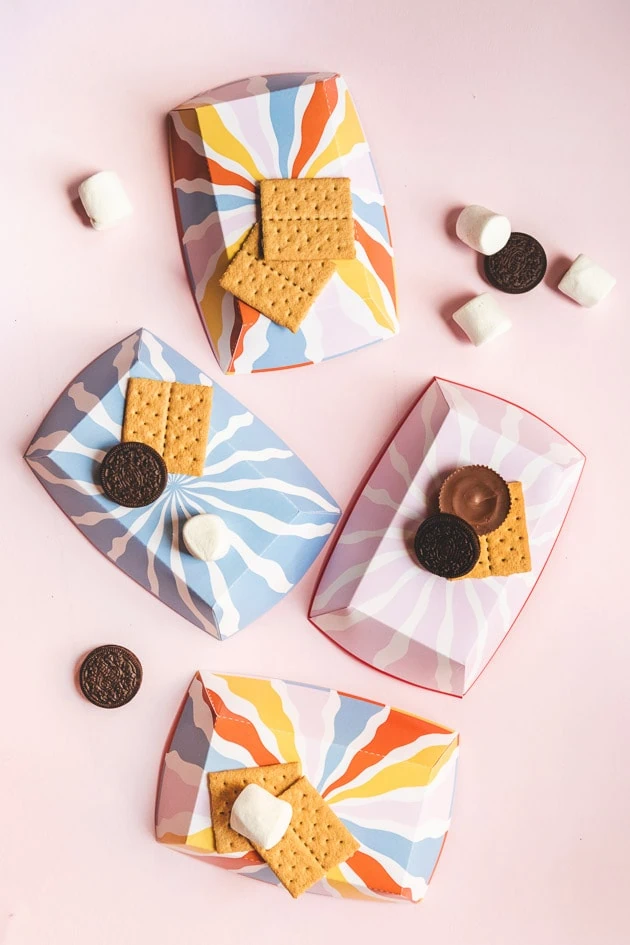 flat lay of blue, pink, and multicolored paper s'more trays on a pink background. Graham crackers, marshmallows, oreos, and peanut butter cups are scattered around. 