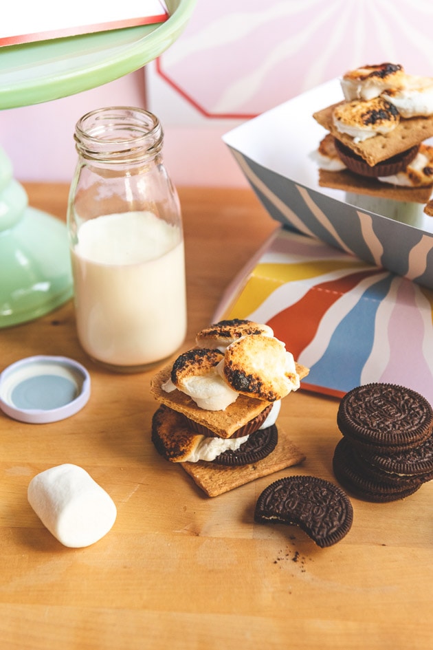 S'mores in colorful paper trays against a pink wall with a jug of milk and some printable templates in the background.
