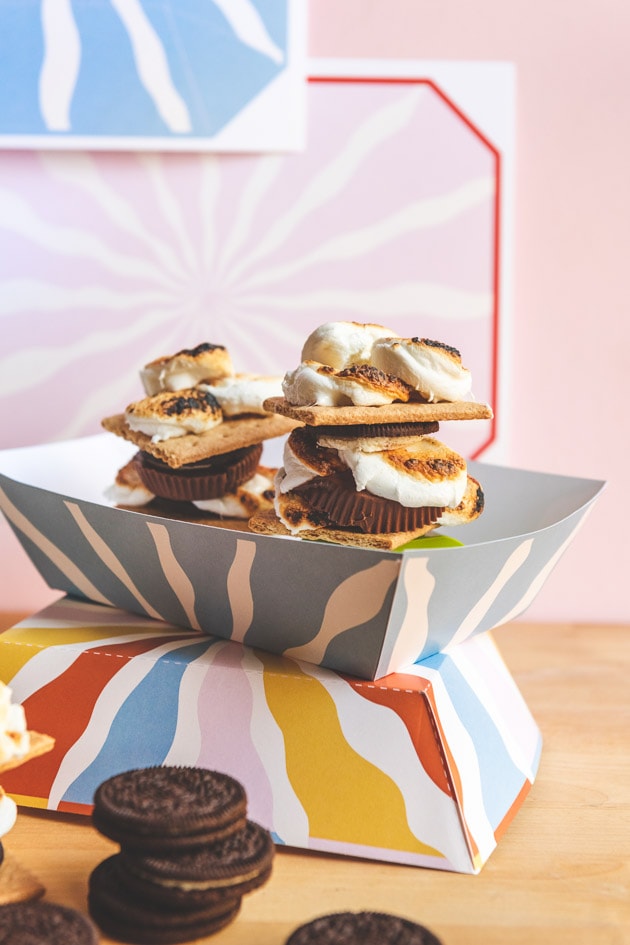 S'mores in colorful paper trays against a pink wall with a jug of milk and some printable templates in the background.