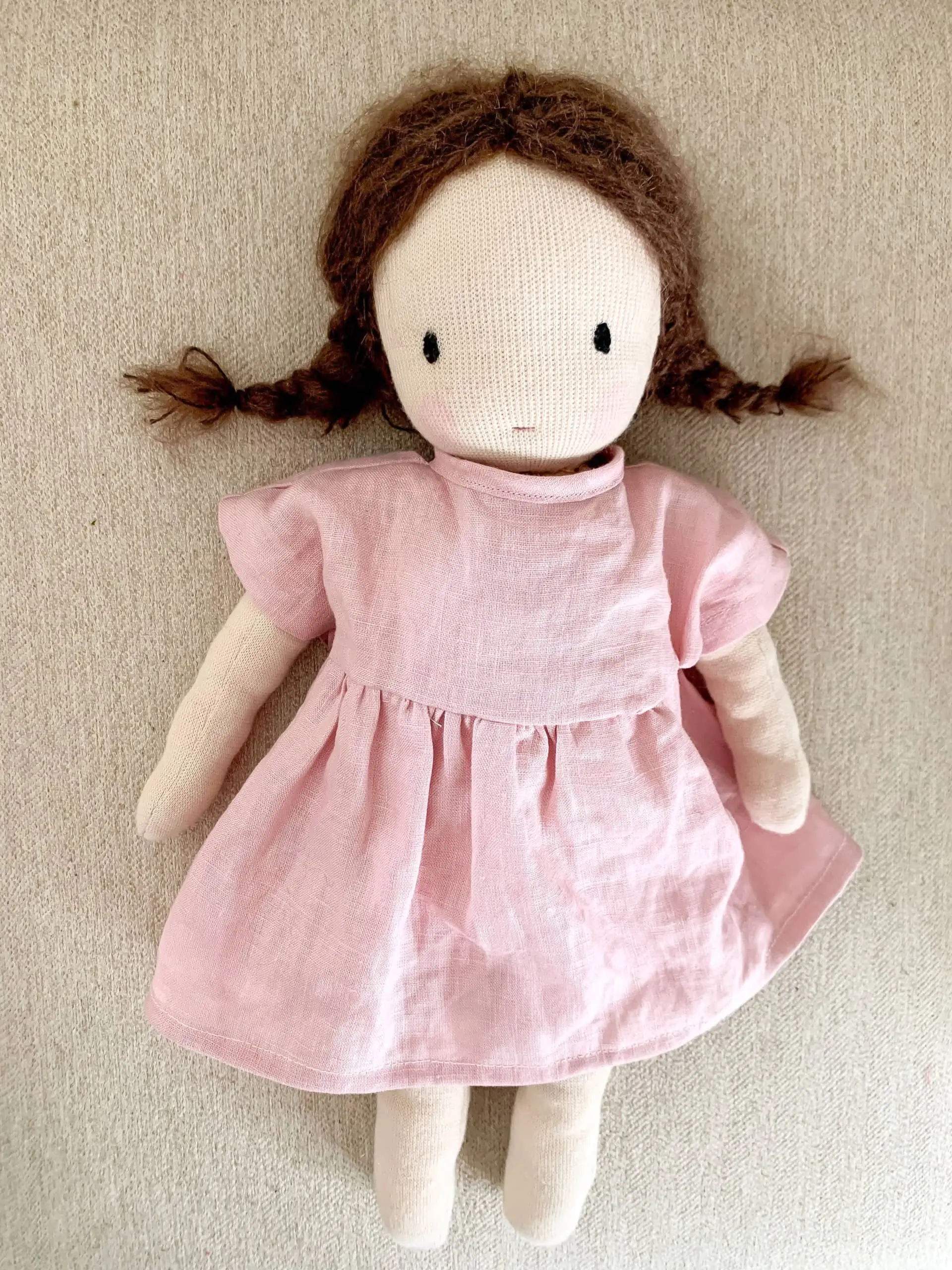 A waldorf-inspired handsewn doll wearing a pink linen dress. The doll has brown braids, pink cheeks, simple features, and is on a beige background.