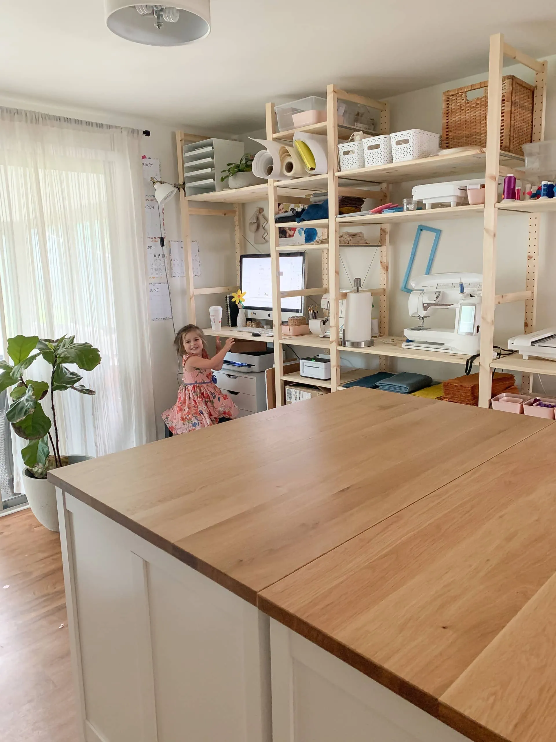 Sarah Cambio's workspace. There's a big wooden island, open wooden shelves filled with materials, and a fiddle leaf fig. Sarah's daughter is wearing pink and sitting by the shelves at a computer.