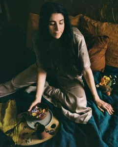 Loria sitting on a bed with teal bedding and flowers in a bowl. She's wearing a beige jumpsuit and there's low, moody lighting.