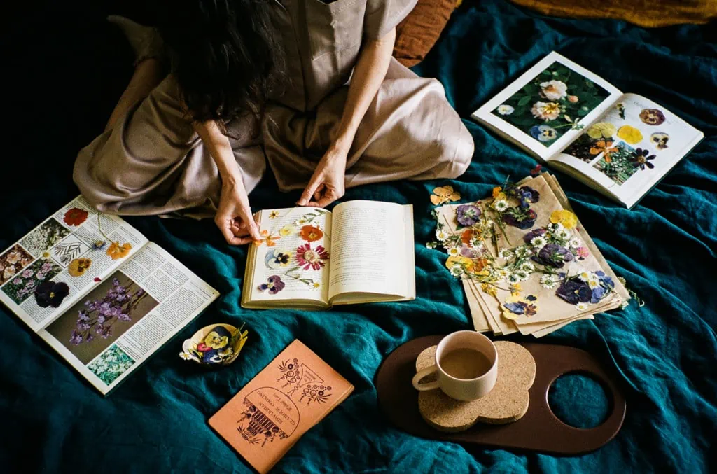 Loria sits cross-legged on a teal blanket surrounded by pressed flowers in books and a cup of tea.