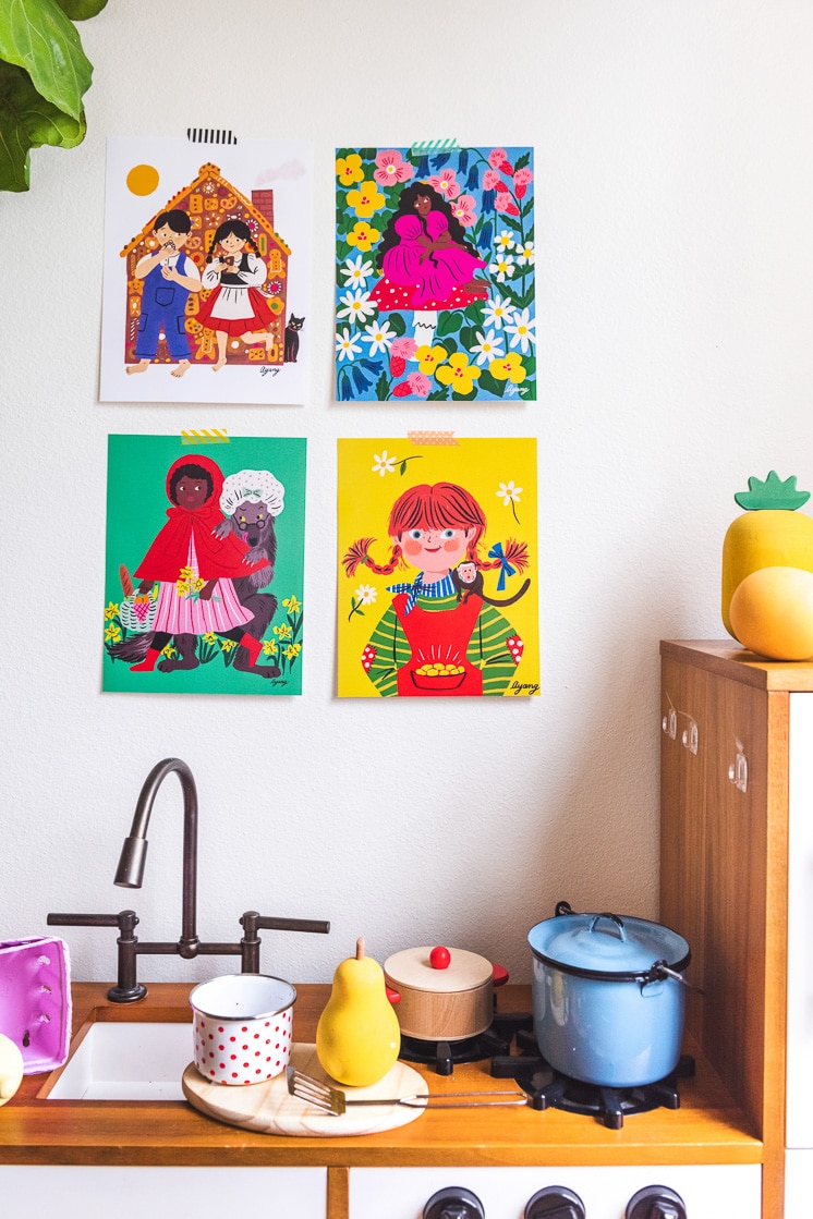 four art prints by Ayang Cempaka hanging above a play kitchen. The prints show hansel and gretel, thumbelina, little red riding hood, and Pippi Longstocking.
