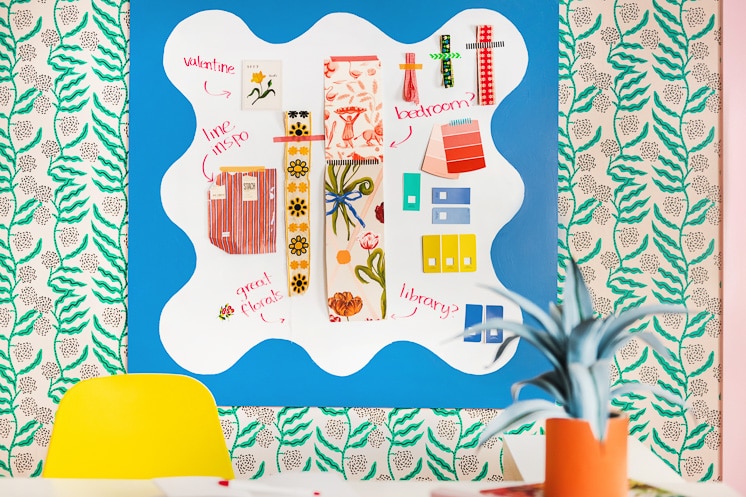 A squiggly-painted whiteboard with paint swatches, ribbons, and other mood board-like objects taped to it. It's on wallpaper with a green botanical design.