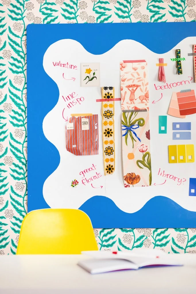 A squiggly-painted whiteboard with paint swatches, ribbons, and other mood board-like objects taped to it. It's on wallpaper with a green botanical design.
