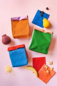 Colorblocked cloth lunch sacks among snacks and wooden fruit on a pink backdrop