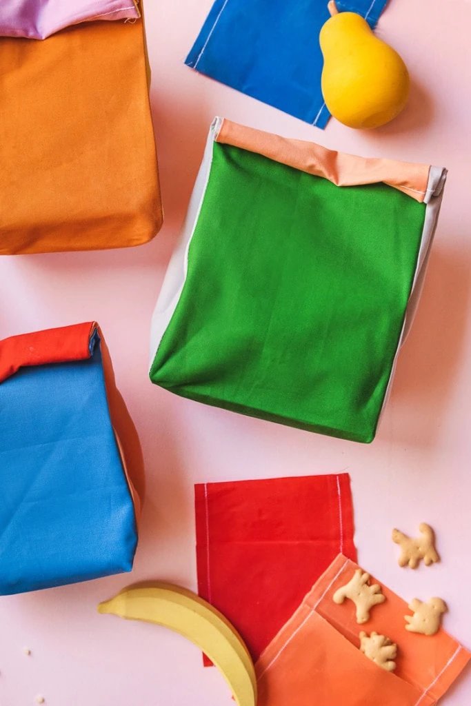 Colorblocked lunch sacks and beeswax snack wraps surrounded by play fruit and animal crackers on a pink background.