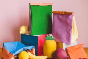 Colorblocked lunch sacks and beeswax snack wraps piled on top of blocks and surrounded by colorful wooden fruit