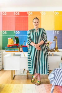 Brittany stands in her rainbow office wearing a green striped dress. Next to her on the desk are some colorblocked reusable lunch sacks.