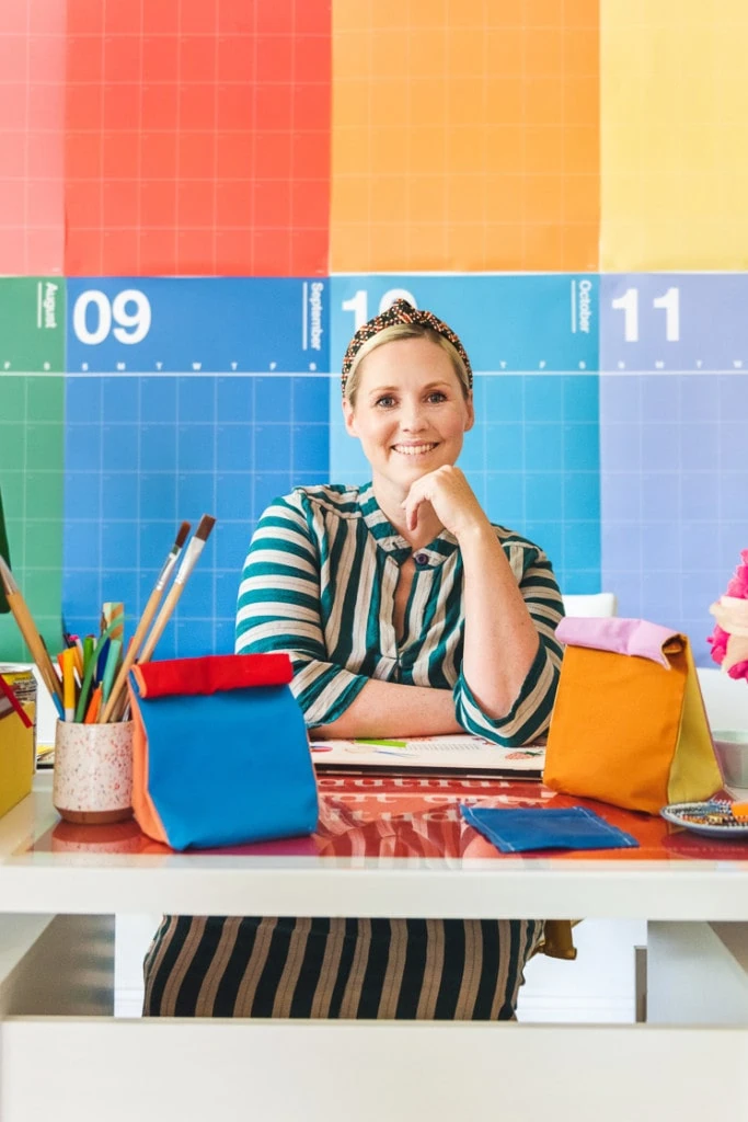 Brittany sits behind her desk surrounded by colorblocked lunch sacks and beeswax snack wraps. THere's a rainbow calendar behind her and she's wearing a striped green dress.