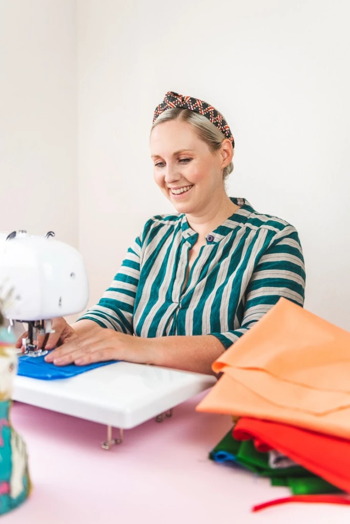 Brittany sits at a sewing machine and makes a beeswax snack wrap. She's wearing a striped green dress and surrounded by colorful, solid fabric.