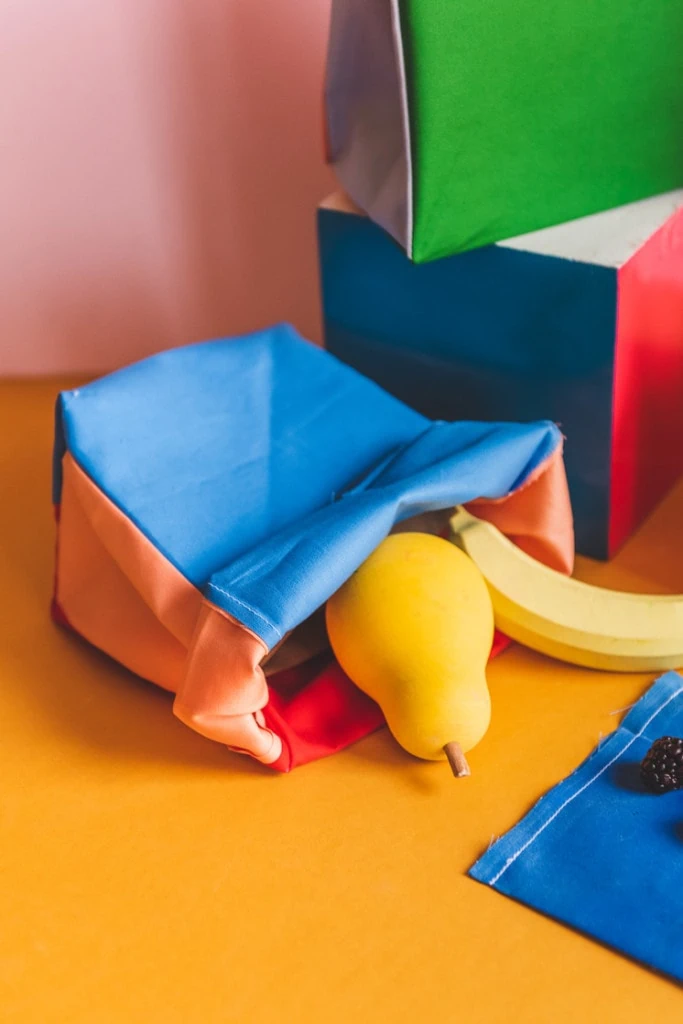 A wooden pear and banana tumble out of a colorblocked lunch sack on a yellow and pink backdrop