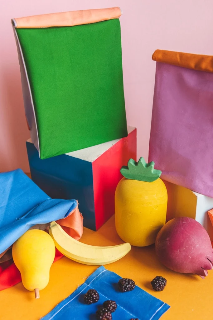 Colorblocked lunch sacks and beeswax snack wraps surrounded by play fruit and blackberries.
