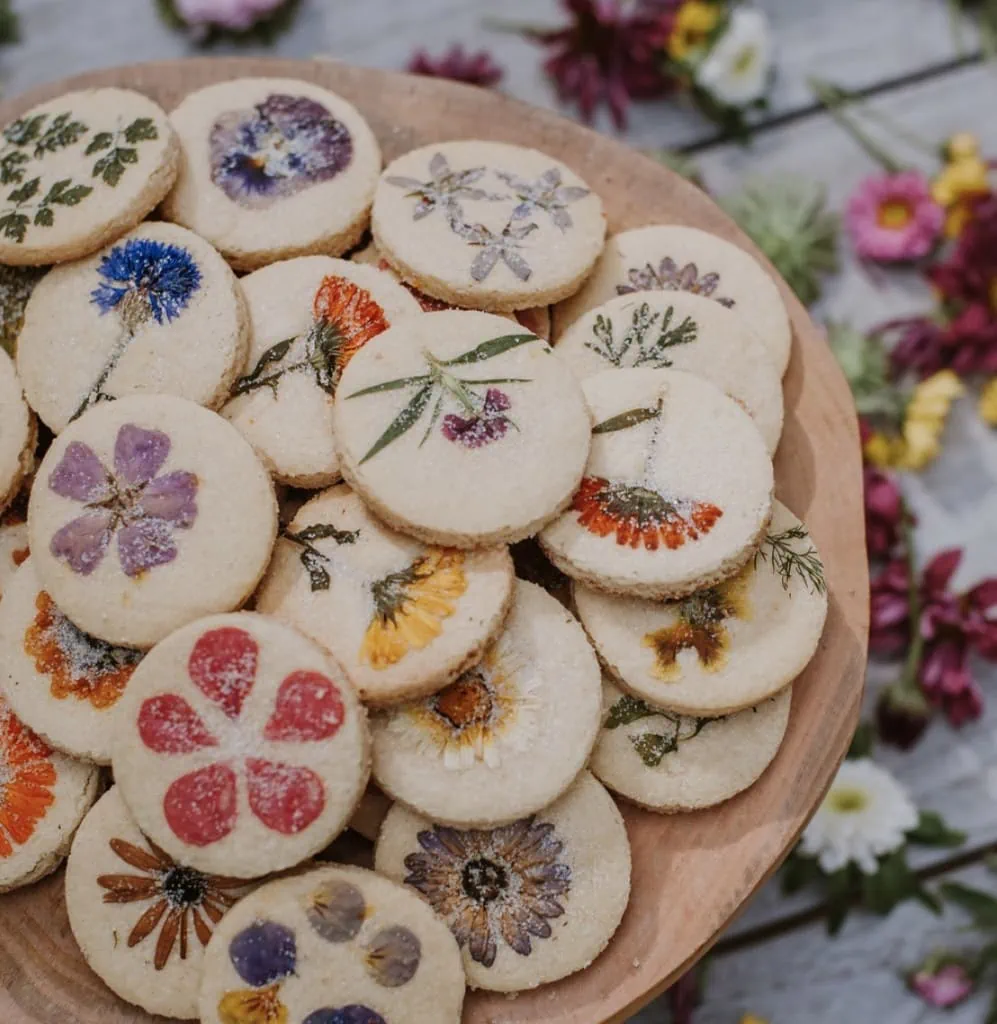 A plate of baked floral shortbread stacked up. In the background there are lots of flowers scattered.