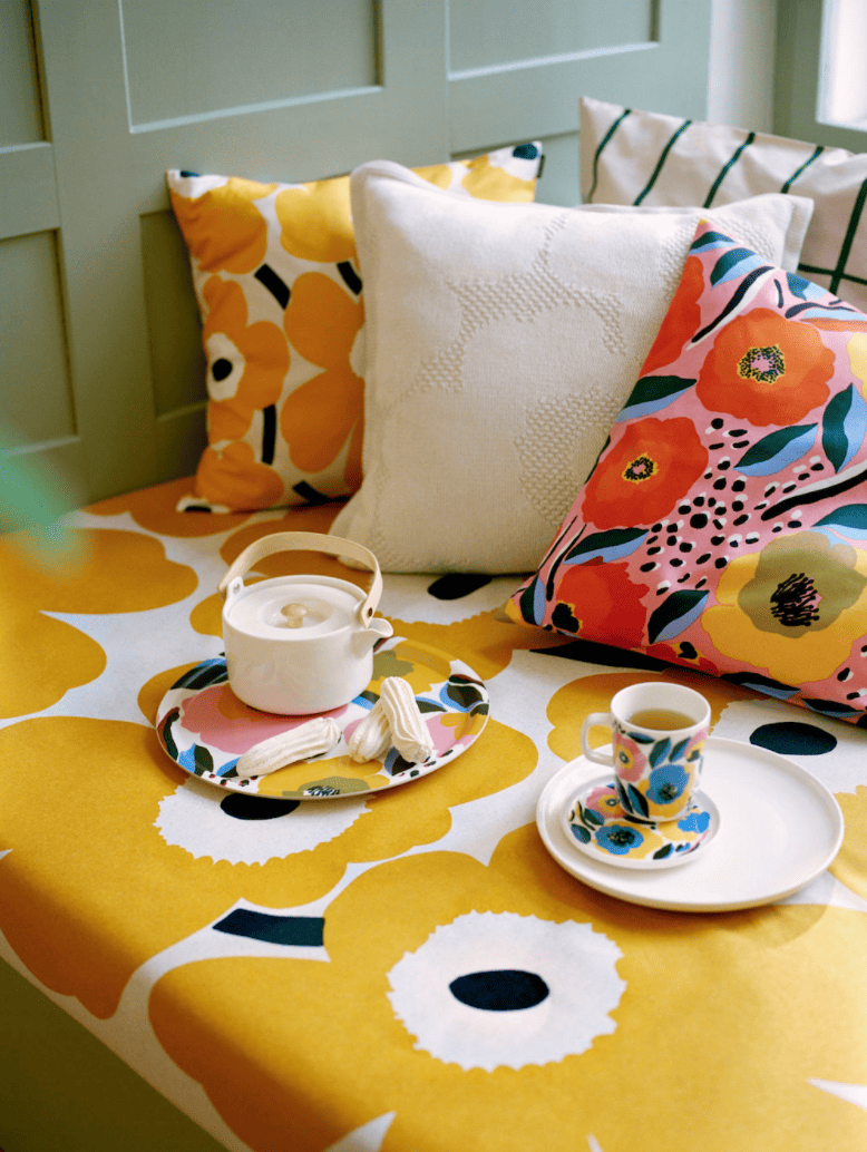 various bright florals on a day bed and as pillows. There is also a pillow with a grid. Tea cups and plates and cookies are set out on the day bed.
