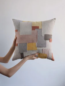 Two arms hold up a patchwork pillow in various shades of blush, gold, and neutrals.