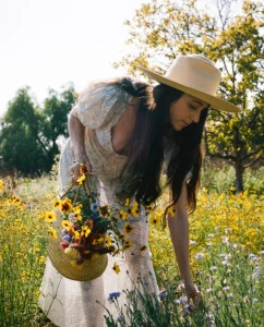 Loria bends down to pick wildflowers in a meadow. She's wearing a white dress and a straw hat.
