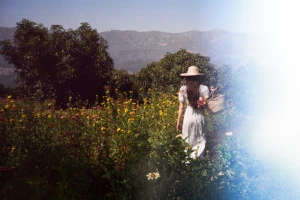A film photograph of Loria walking away from the camera through a field of wildflowers. She's wearing a white dress and a straw hat and there are trees and mountains in the background.