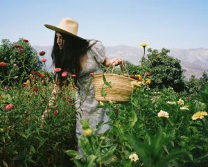 Loria stands in a field of zinnias wearing a white dress and a straw hat. She's holding a basket full of flowers.