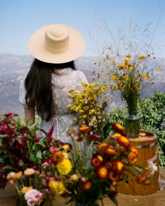 Loria stands with her back to the camera. She's surrounded by fresh cut flowers and she's wearing a straw hat, and there are misty mountains in the background.