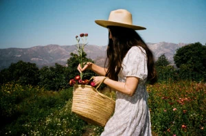 Loria walking through a meadow of flowers wearing a white dress and a straw sun hat. She's holding a basket of flowers and the sky is blue.
