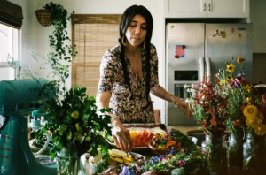 Loria is wearing a floral dress and standing in a kitchen surrounded by colorful produce.