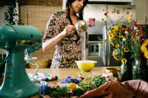 Horizontal photo of Loria measuring sugar into a yellow mixing bowl. She's in a kitchen and surrounded by flowers and a turquoise kitchenade mixer