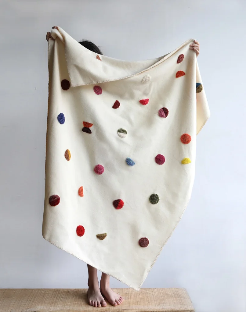 Someone stands on a bench holding a white punch needle blanket above their head. The punch needled parts are in lots of colors and look like confetti dots sprinkled throughout the blanket's surface.